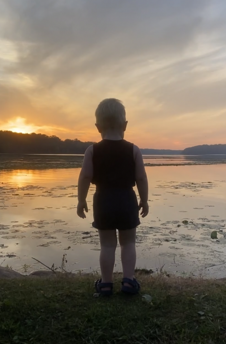 A small child stands on the edge of a lake while looking out at the sunset.