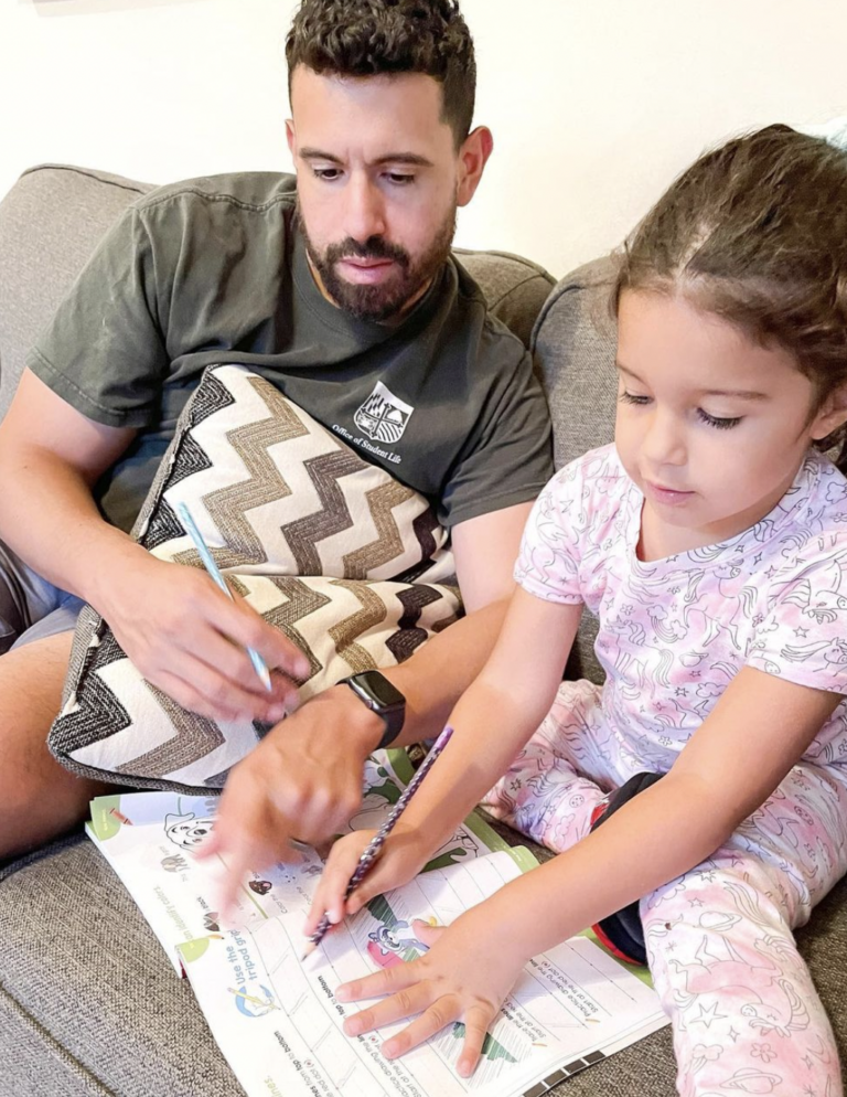 Man helping his daughter with a workbook while seated on the couch.