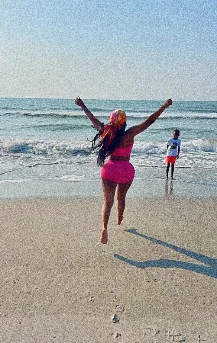 A woman runs toward ocean waves with hands extended towards the sky while a young boy watches close by.