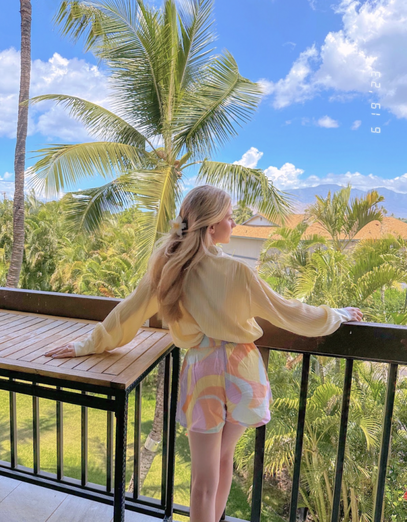 A woman looks out at palm trees while standing on a balcony.