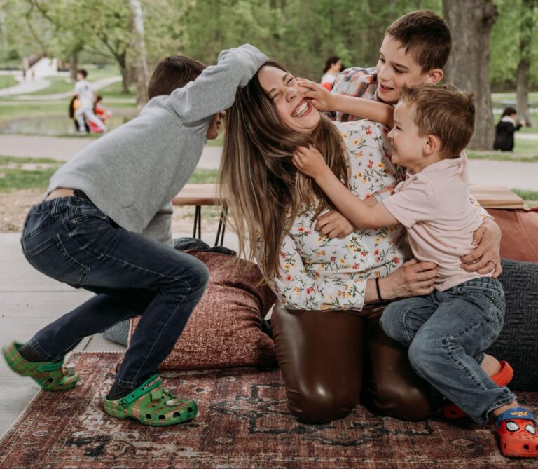 A woman smiles while surrounded by her three sons hugging and playing with her.