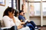 People sit in doctor's waiting room.  One woman looks a medical brochure and a man uses his smart phone to make a call.